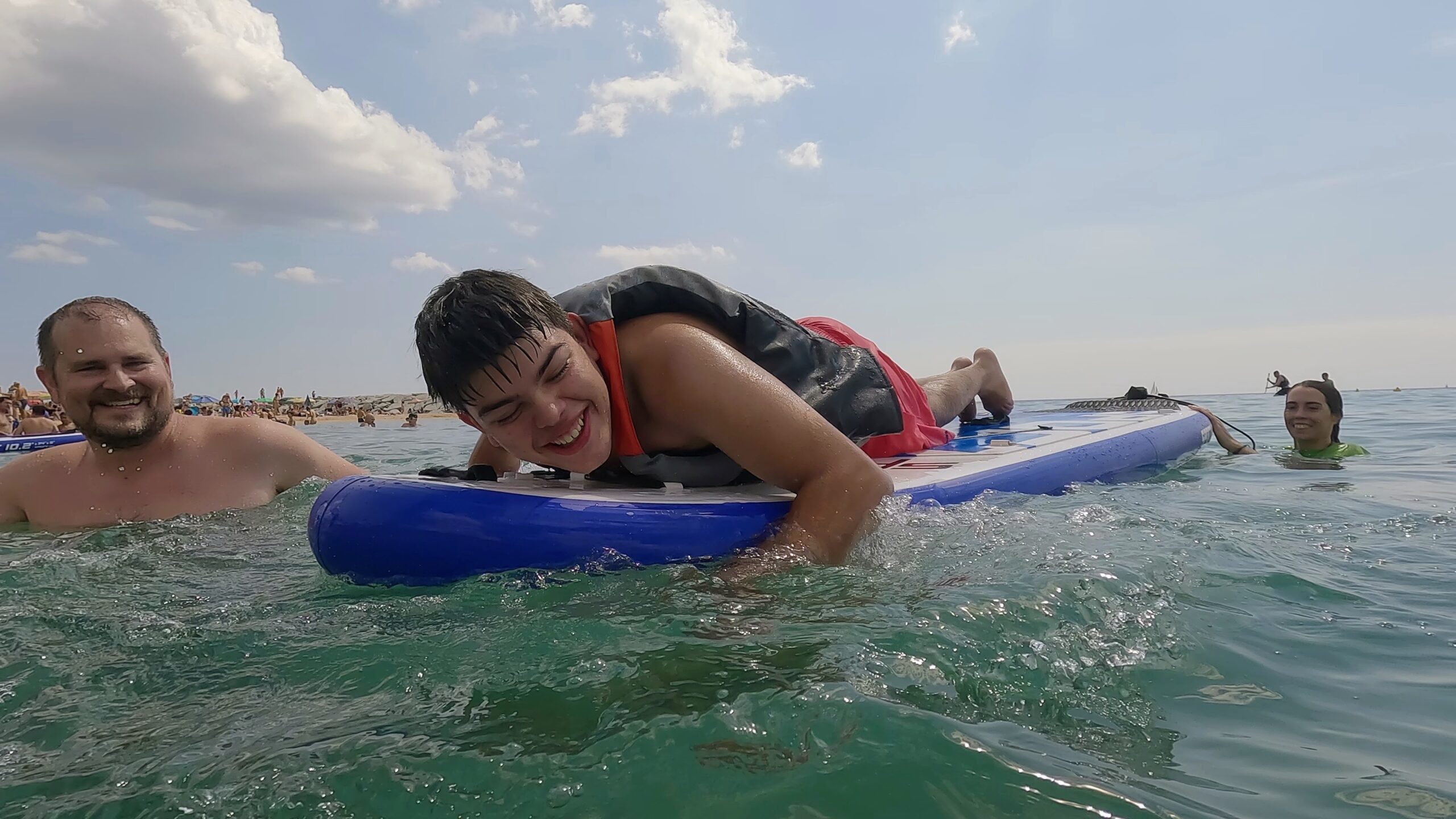 Joven disfrutando sobre una tabla de paddle surf adaptado en la playa de Calonge i Sant Antoni durante Onades Inclusives 2025, acompañado por voluntariado en el agua.