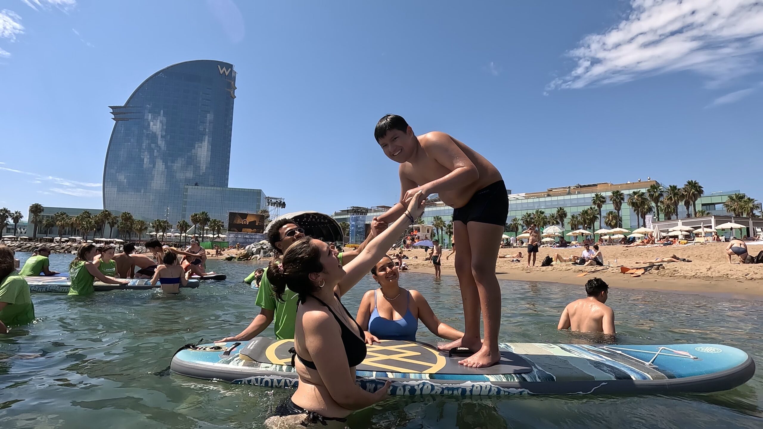 Niños y voluntarios disfrutando del surf inclusivo en la playa de Sant Sebastià durante una actividad de SUPerando con Acordis, SeaYou e IAU Barcelona.