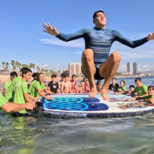 Alumno de la Escola REL disfrutando de una sesión de surf inclusivo con SUPerando en la playa de Sant Sebastià, junto al equipo de SeaYou y estudiantes del Institut de Nàutica de Barcelona