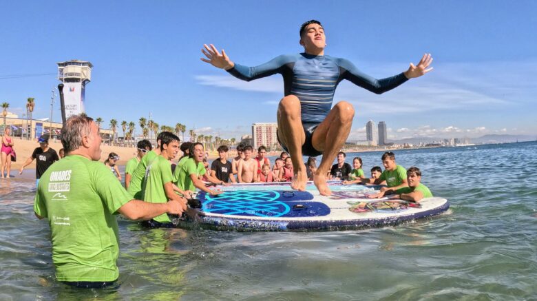 Alumno de la Escola REL disfrutando de una sesión de surf inclusivo con SUPerando en la playa de Sant Sebastià, junto al equipo de SeaYou y estudiantes del Institut de Nàutica de Barcelona